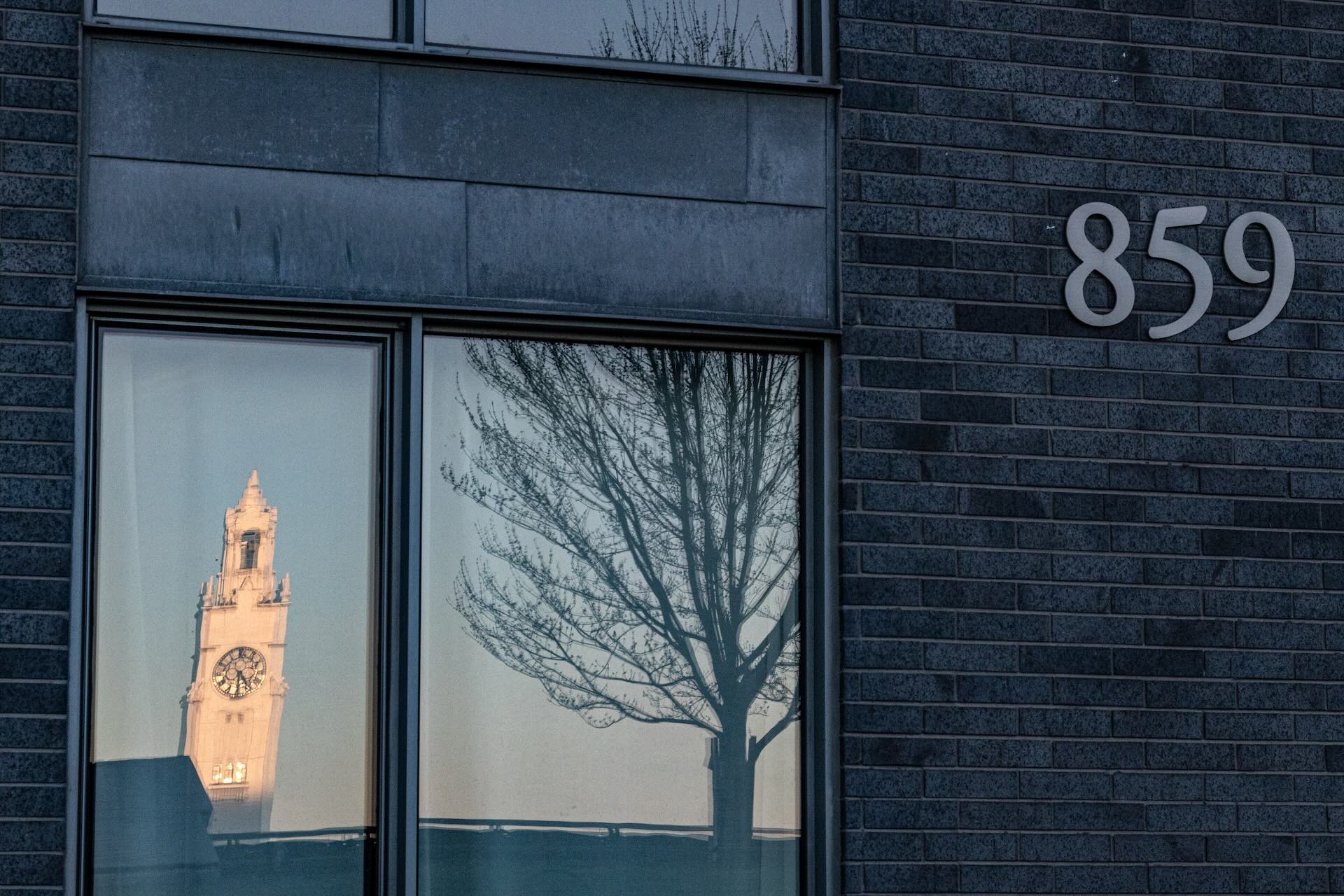 Reflection of a clock tower and a tree in a window of a dark brick building with the number 859. The scene conveys a serene, contemplative tone.