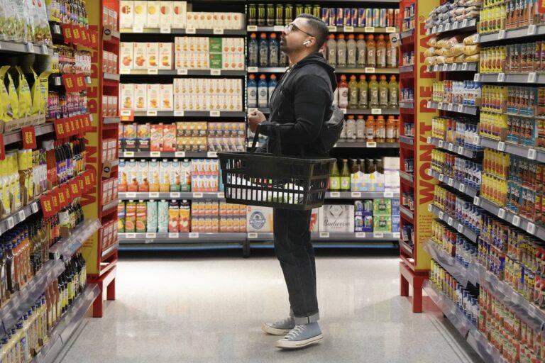A man in a black outfit stands in a brightly lit grocery aisle, holding a shopping basket. Shelves filled with various products line both sides.