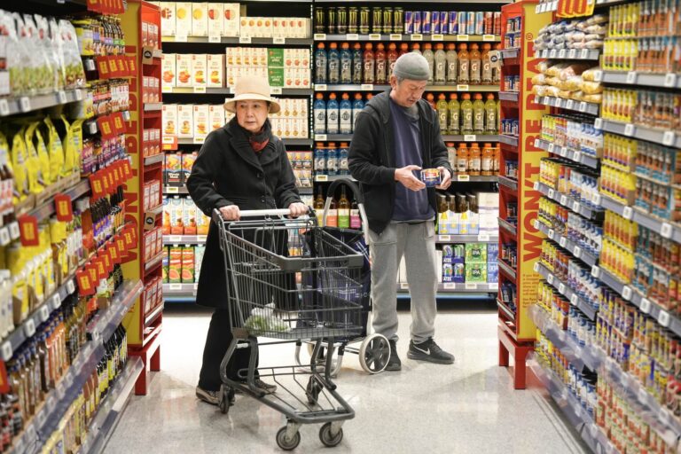 Two people are seen shopping in the aisles of a No Frills grocery store. One person is pushing a shopping cart and the other is examining a product.
