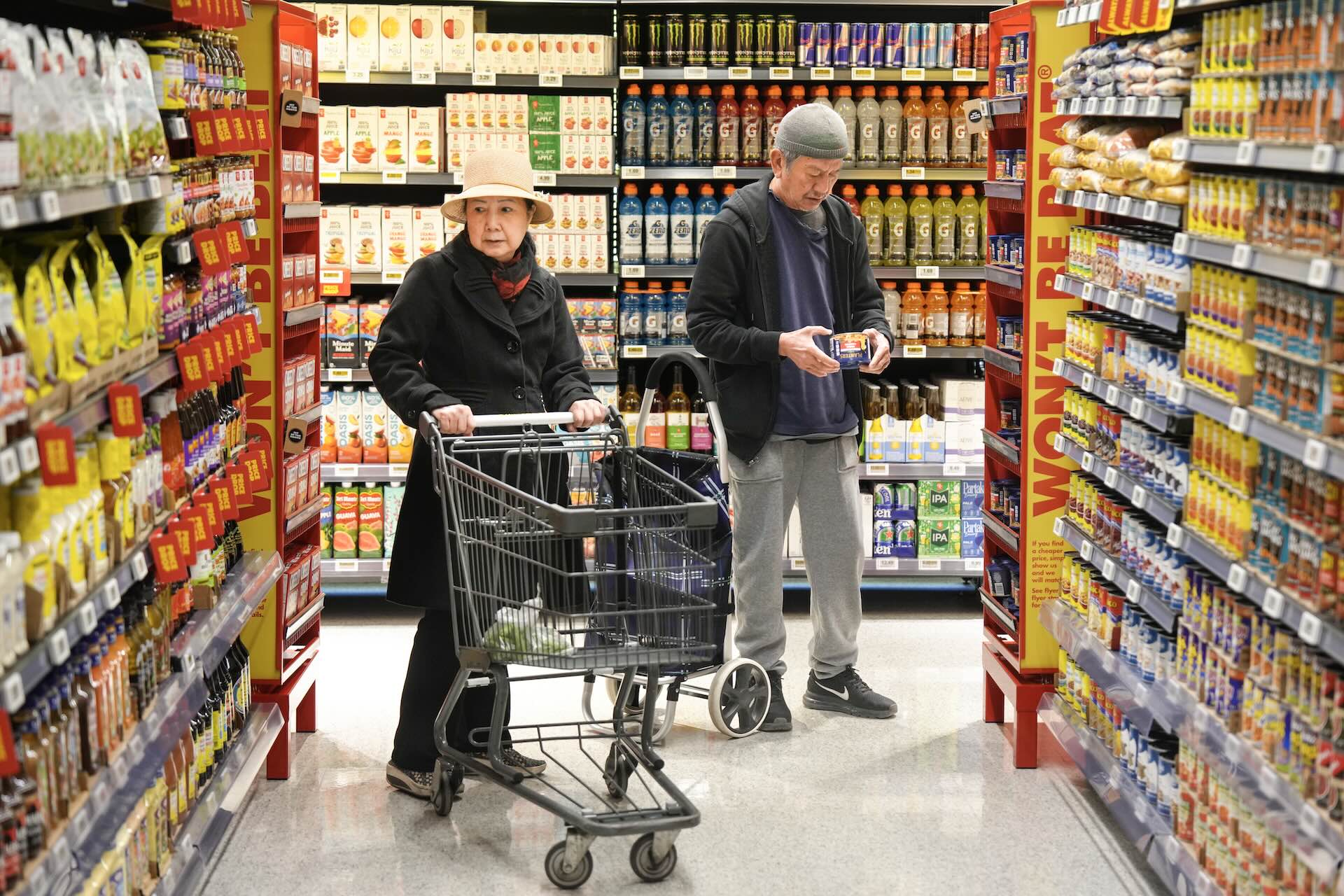 Two people are seen shopping in the aisles of a No Frills grocery store. One person is pushing a shopping cart and the other is examining a product.