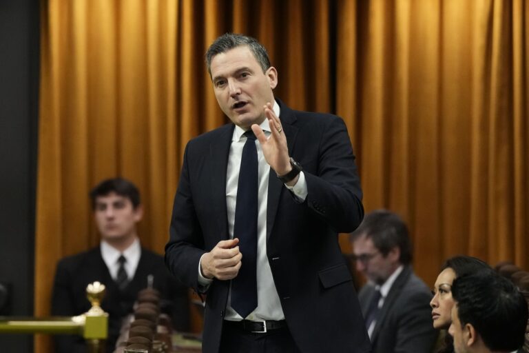 Evan Solomon in a suit and tie, gesturing with his left hand as he speaks, Several people sit and stand behind him looking in other directions. There's an orange curtain behind him lit from above.