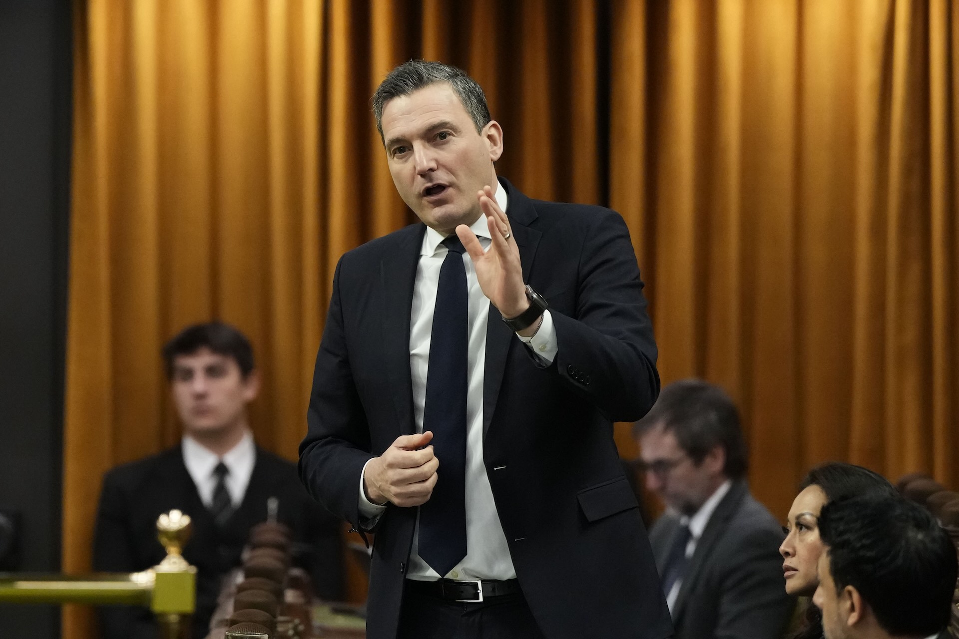 Evan Solomon in a suit and tie, gesturing with his left hand as he speaks, Several people sit and stand behind him looking in other directions. There's an orange curtain behind him lit from above.