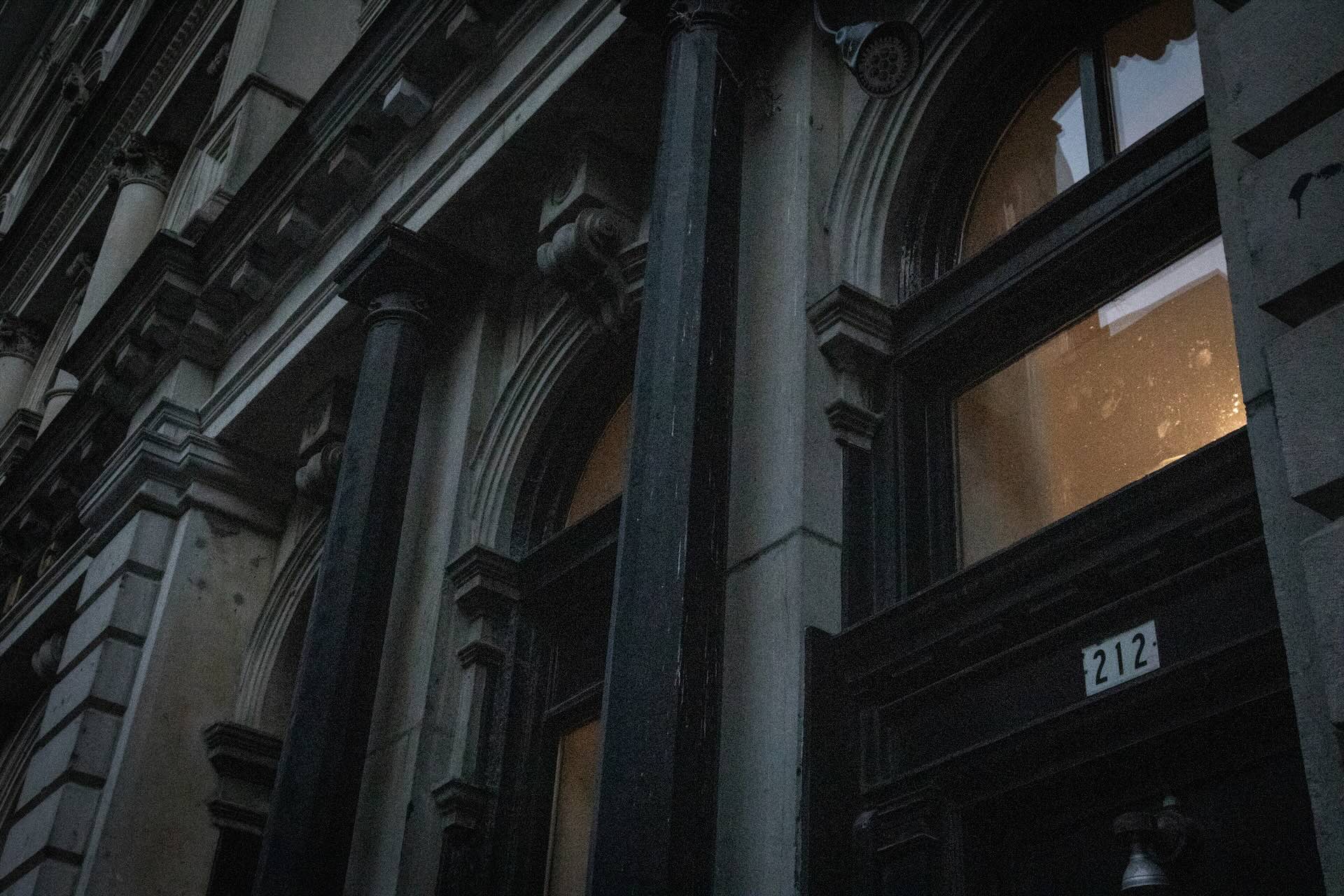 A dimly lit, historic building facade with ornate columns and arched windows. A small lit room is visible through a window, creating a mysterious ambiance.