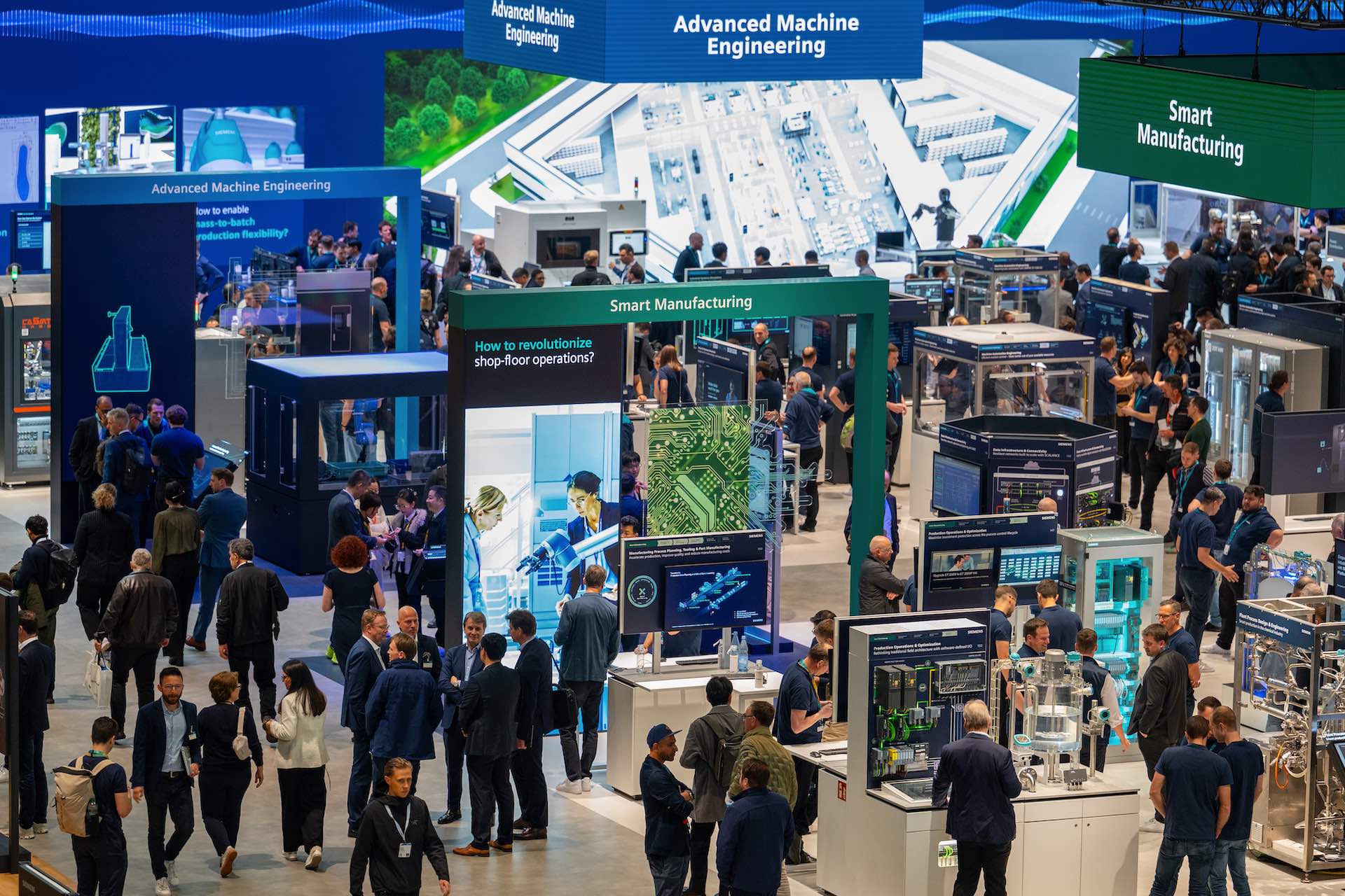 A high-angle shot of one of the pavilions at Hannover Messe, showing people lingering at a variety of robotics and technological display booths. Signs reading "Advanced Machine Engineering" and "Smart Manufacturing" hang from the ceiling.