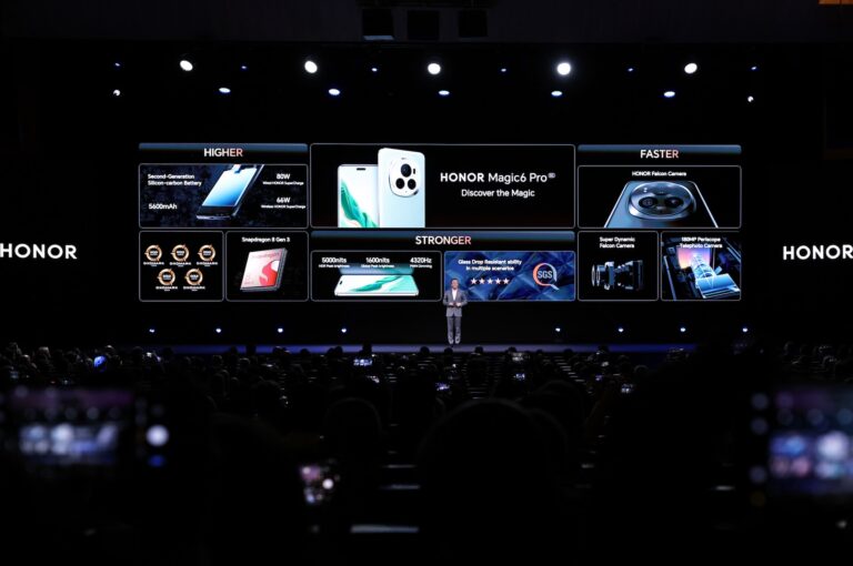 A wide shot of a man in a grey suit on a dark stage, with images of Honor Device products projected behind him.