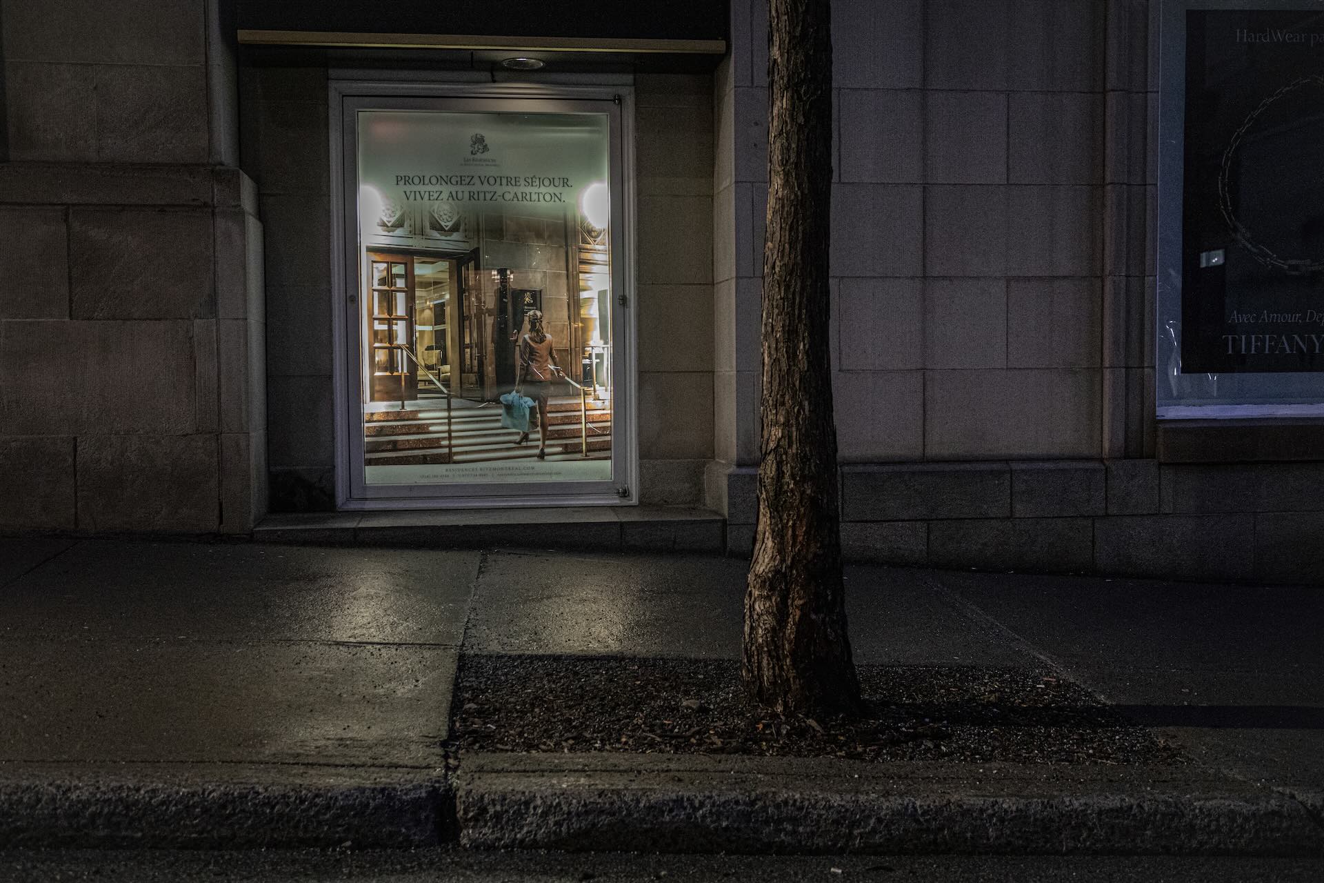 Dimly lit street with a tree and an illuminated display window showing a woman in a dress and heels on a staircase. Text promotes the Ritz-Carlton.