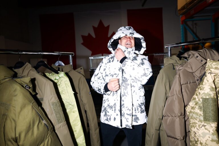 A shot of a laughing James Yurichuk bundling himself up in a white-and-grey camouflage parka, surrounded by other khaki and camouflage parkas on racks. There is a Canadian flag visible on the wall in the darkened background.