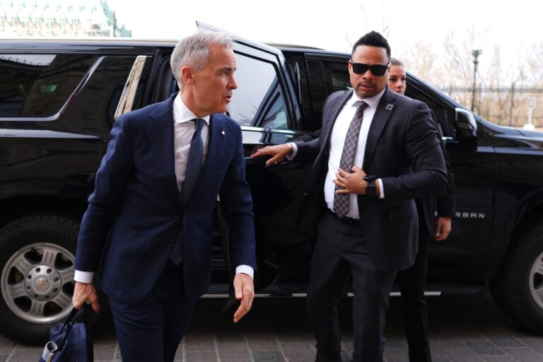 Mark Carney emerges from a black van. A man wearing a dark suit and sunglasses stands behind him.