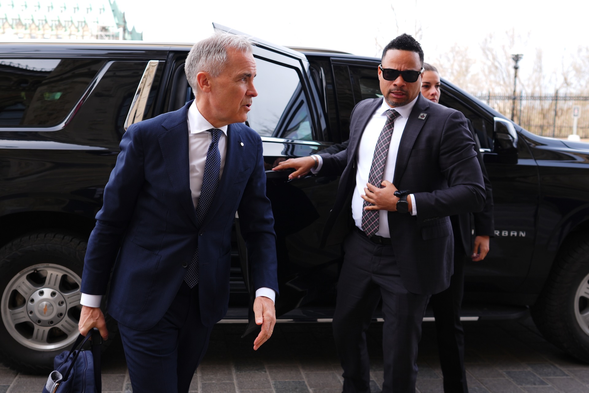 Mark Carney emerges from a black van. A man wearing a dark suit and sunglasses stands behind him.