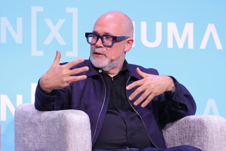 A man wearing glasses and a dark buttoned-down shirt speaks onstage at an event. The background is blue and he sits in an armchair.