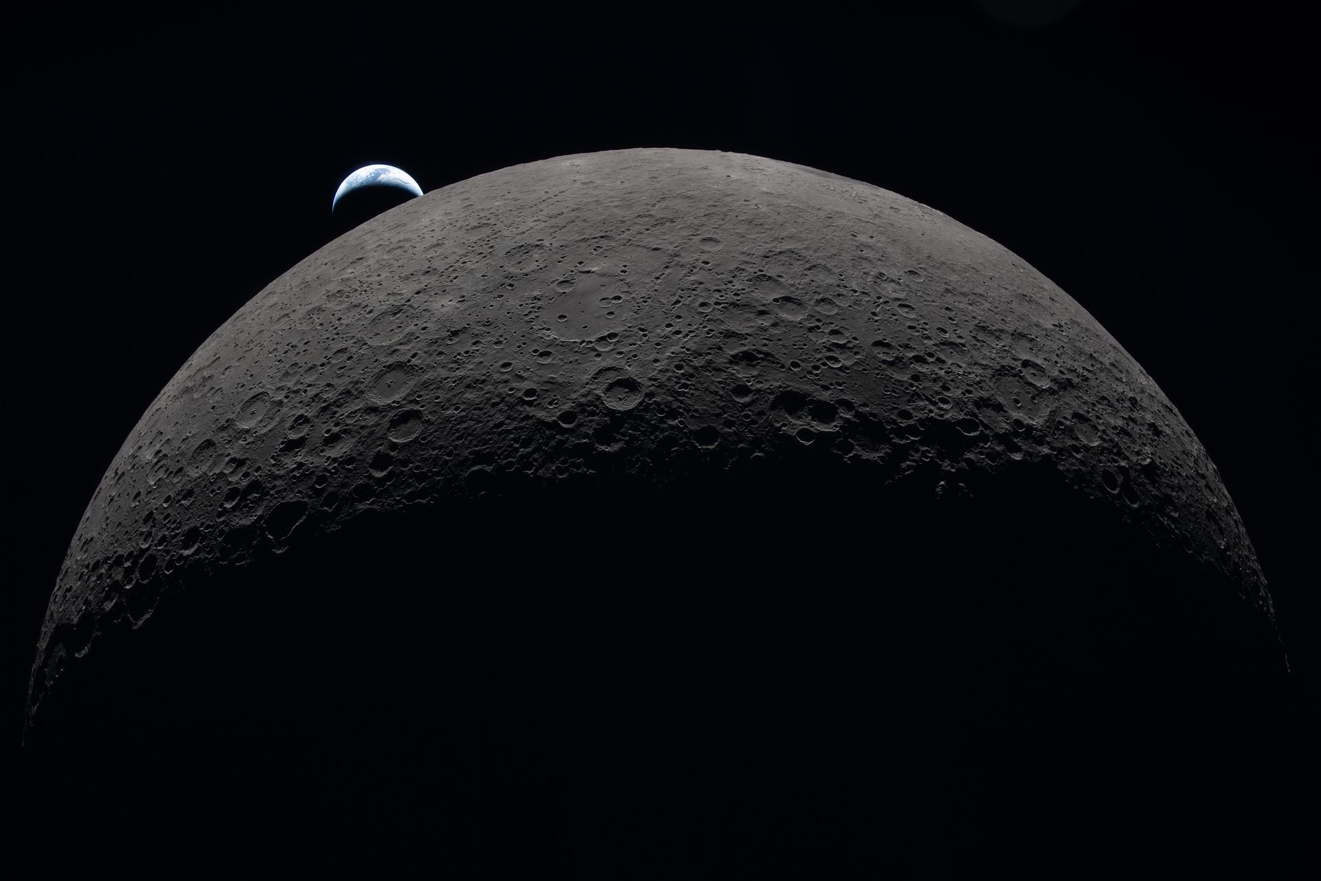 A photo dominated by about two thirds of the moon in the foreground, with Earth appearing much smaller in the background as a mere crescent of blue and white light. Earth appears to be setting on the sharply curved horizon of the moon.