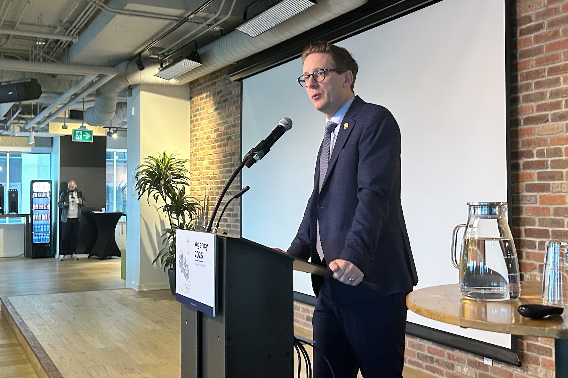 A shot of Nate Glubish at a lectern, against a backdrop of exposed brick partly covered by a white film screen.