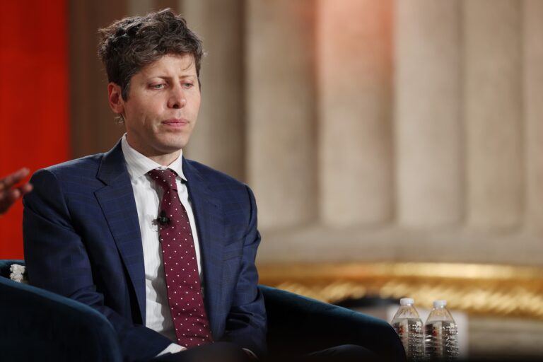 Sam Altman sits on a chair onstage in a navy blue suit and red tie. He has ruffled grey-brown hair and looks pensively at the ground.