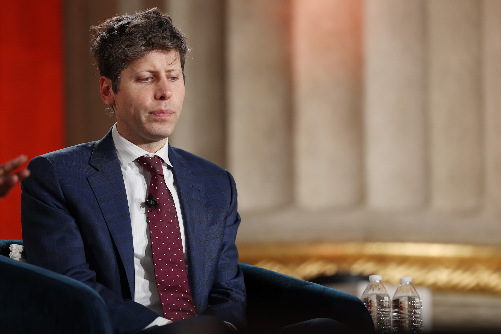 Sam Altman sits on a chair onstage in a navy blue suit and red tie. He has ruffled grey-brown hair and looks pensively at the ground.