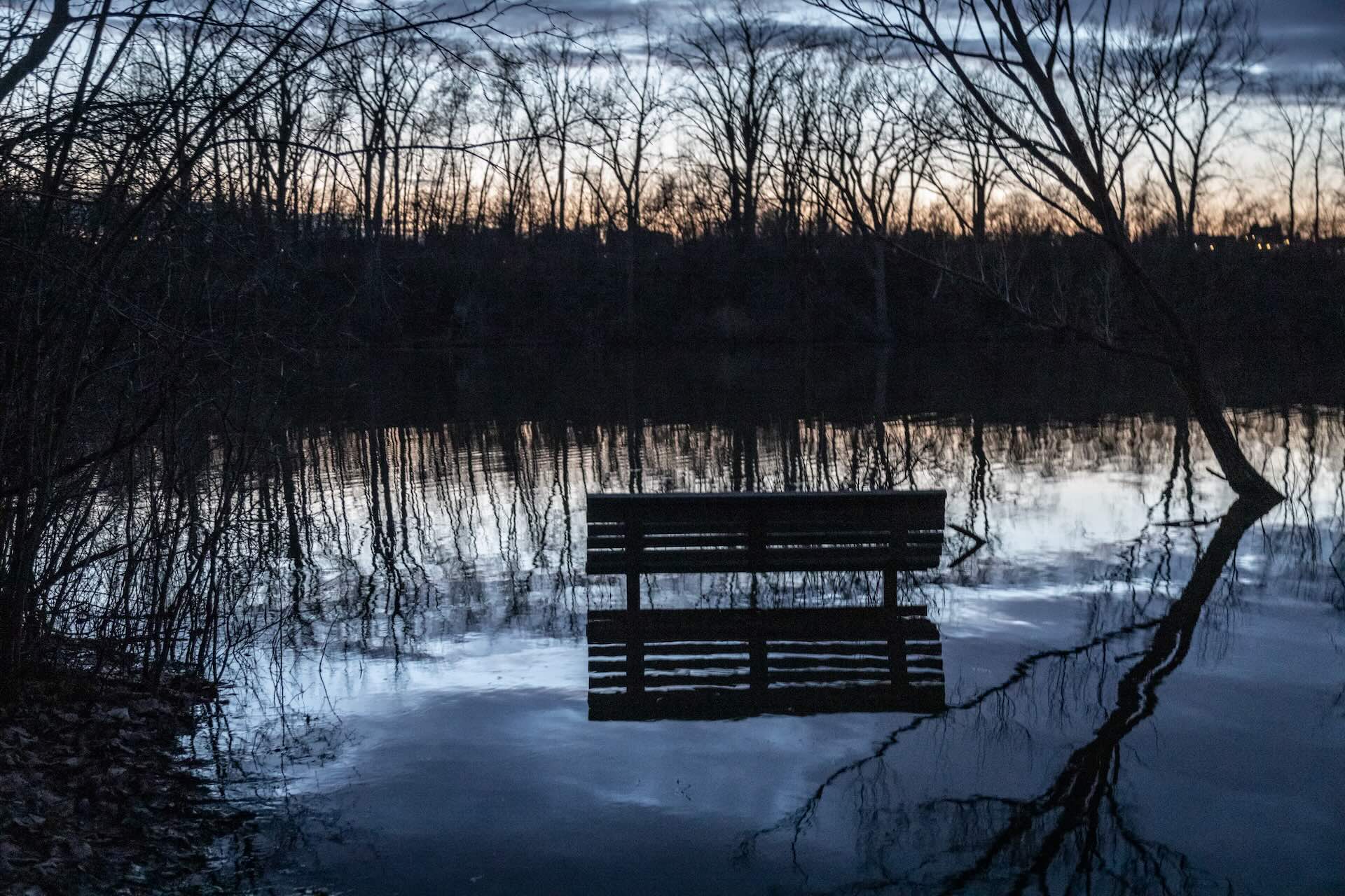 A submerged park bench is reflected in a calm, flooded area at dusk. Bare trees and a fading horizon create a serene, melancholic atmosphere.