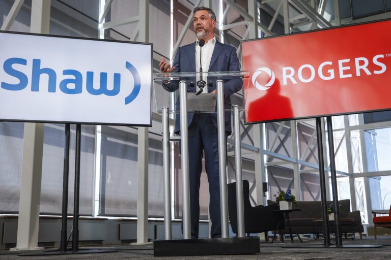 A shot of Rogers CTO Jorge Fernandes standing at a lectern between two screens—one showing the Rogers logo; the other showing the Shaw logo.