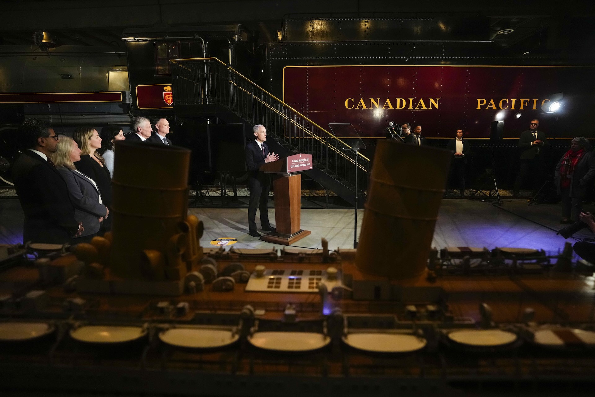 A wide shot of Mark Carney at a lectern, with a vintage Canadian Pacific steam locomotive as his backdrop.