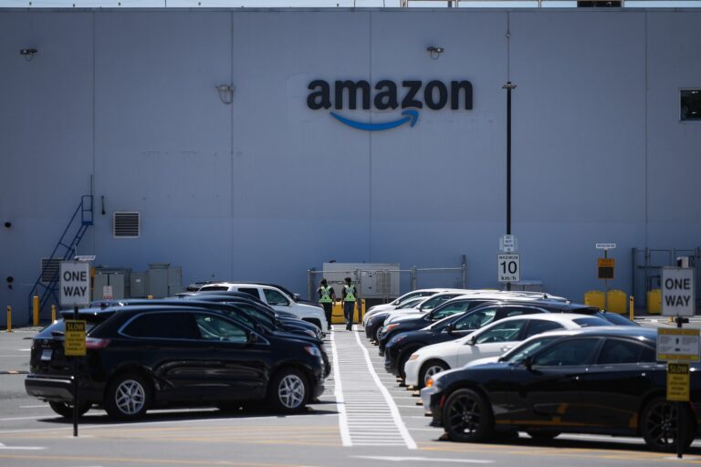 An image of the side of a building with the Amazon logo on it. A parking lot full of cars is visible in the foreground.