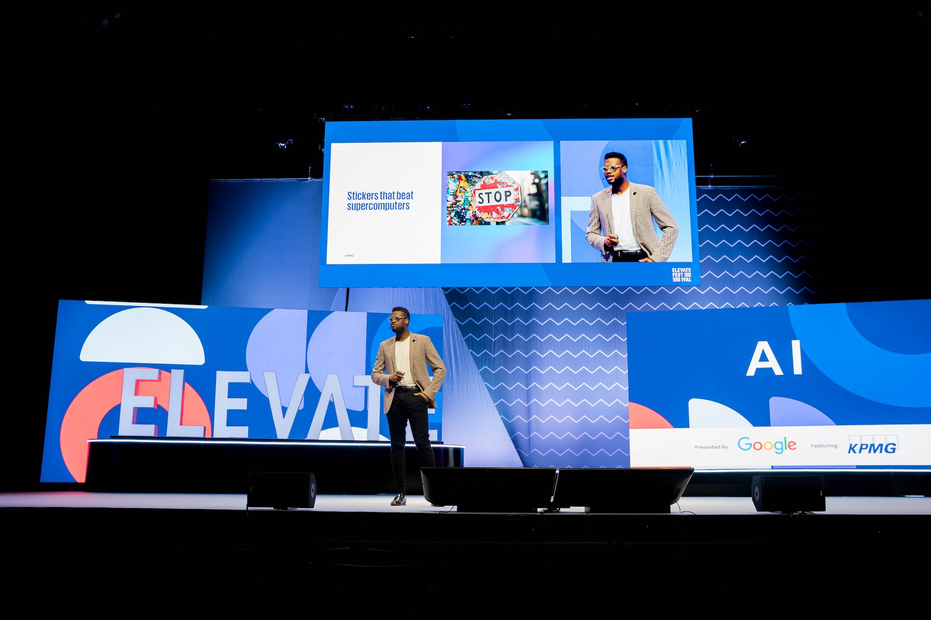 Andrew Forde, wearing a beige tweed blazer, black slacks and a white sweater, speaks on a stage at the Elevate conference in Toronto with three large blue screens in the backdrop. One screen displays the session topic, AI, another displays the logos for sponsors KPMG and Google, and a third screen depicts a photo of a stop sign covered in stickers. The stop-sign photo is labelled, “Stickers that beat supercomputers.”