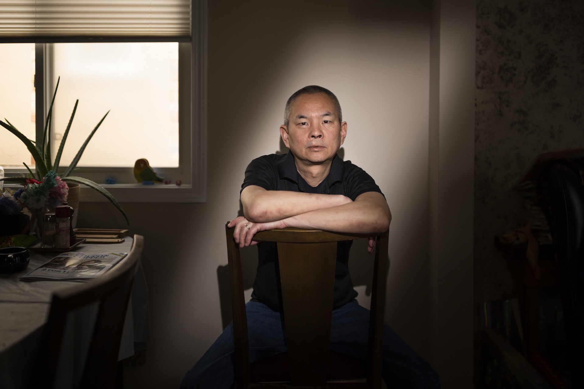 A head-on shot of Anthony Hu looking over the back of a dining chair with his arms folded in front of him. He is in a semi-dark room but is illuminated by a bar of light.