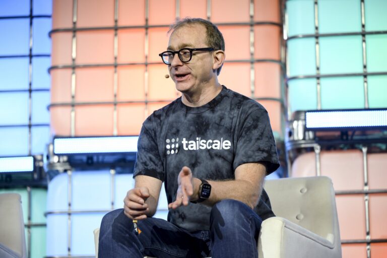 A shot of Avery Pennarun on stage in front of a colourful backdrop. He's wearing a grey t-shirt with Tailscale's logo on it.