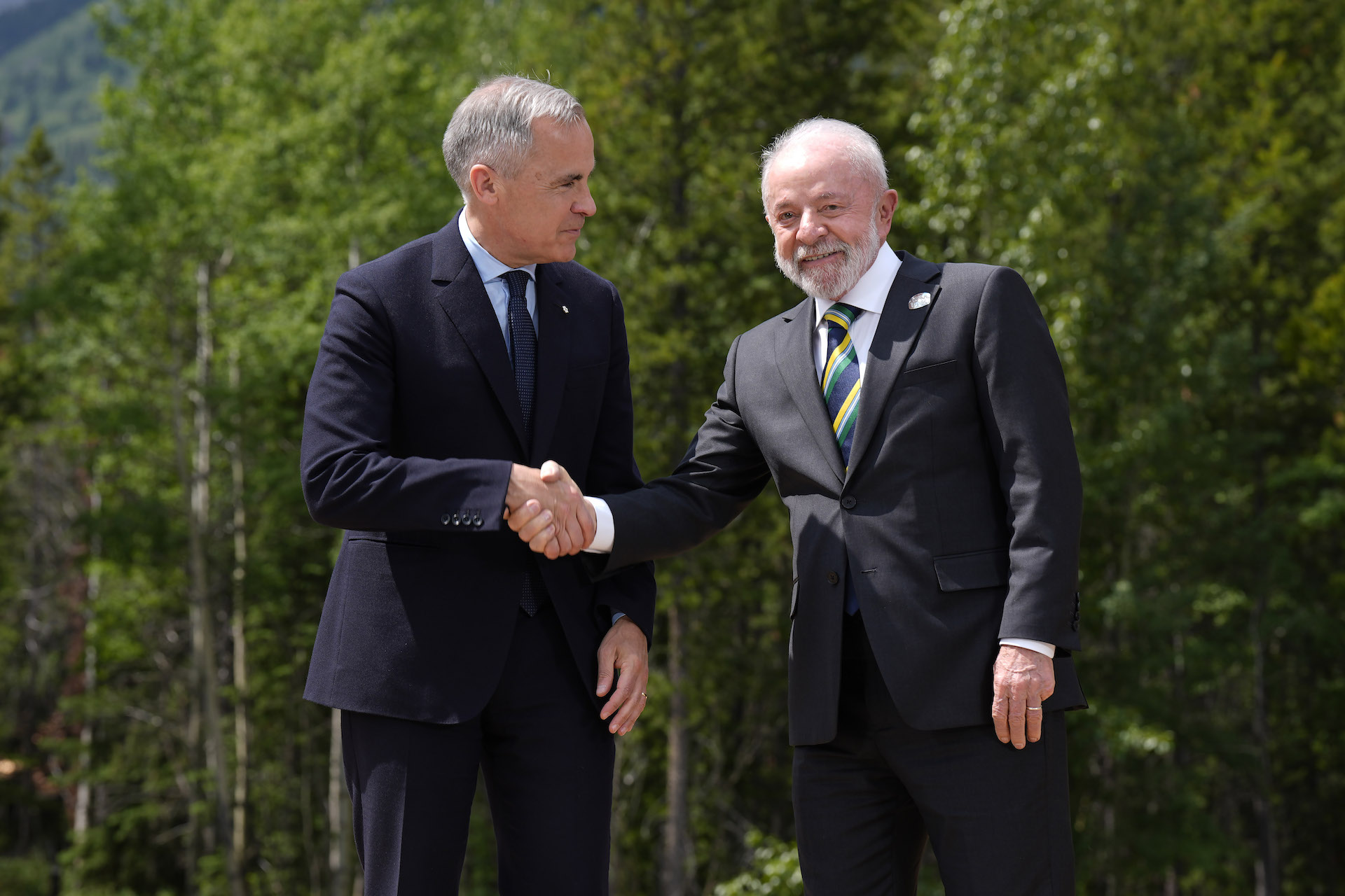 A shot of Mark Carney shaking hands with Lula, the Brazilian president, against a backdrop of trees and mountains.