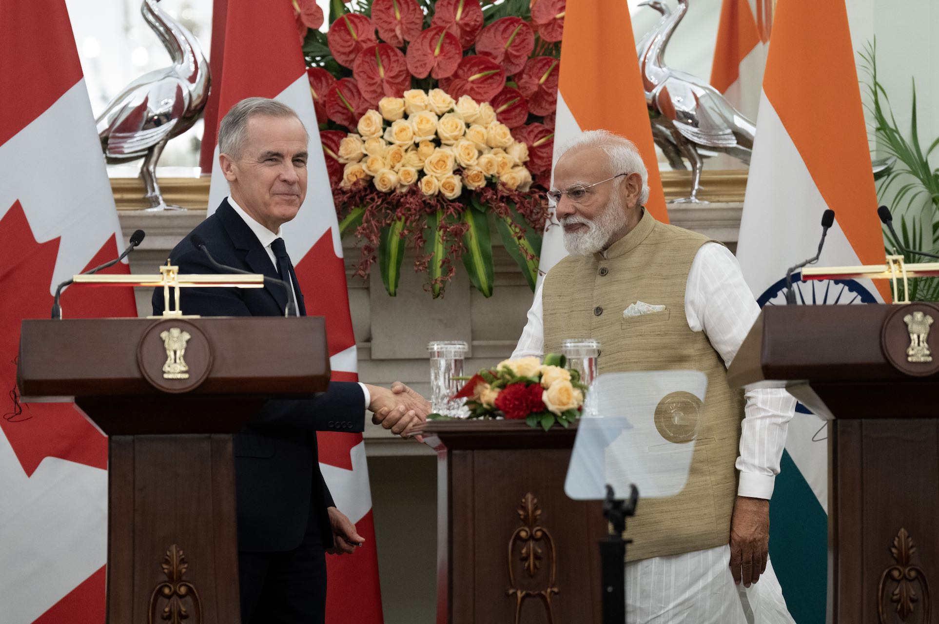 Mark Carney shaking hands with Narendra Modi behind a pair of lecterns. There are Canadian and Indian flags in the background, along with a large floral display.