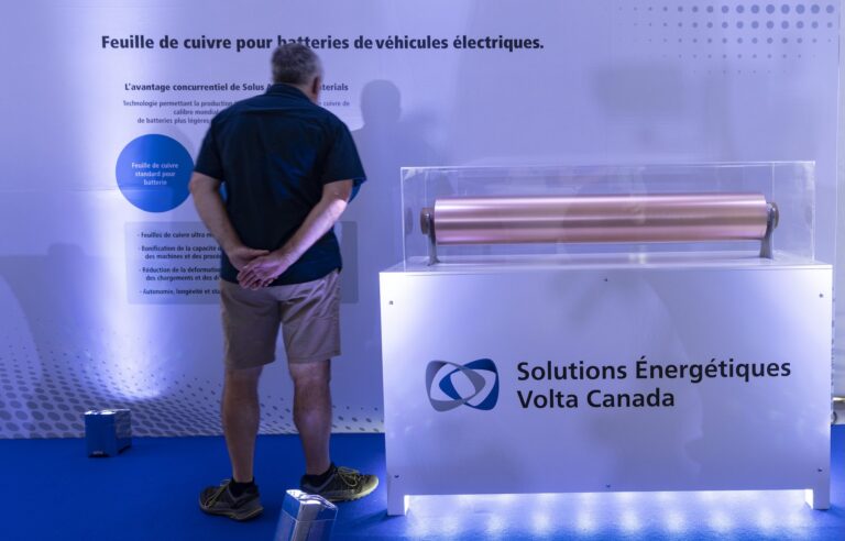A man wearing a dark t-shirt and beige shorts stands facing a display of a sample roll of copper foil, visible in the foreground. A sign below the foil reads Solutions énergétiques Volta Canada.
