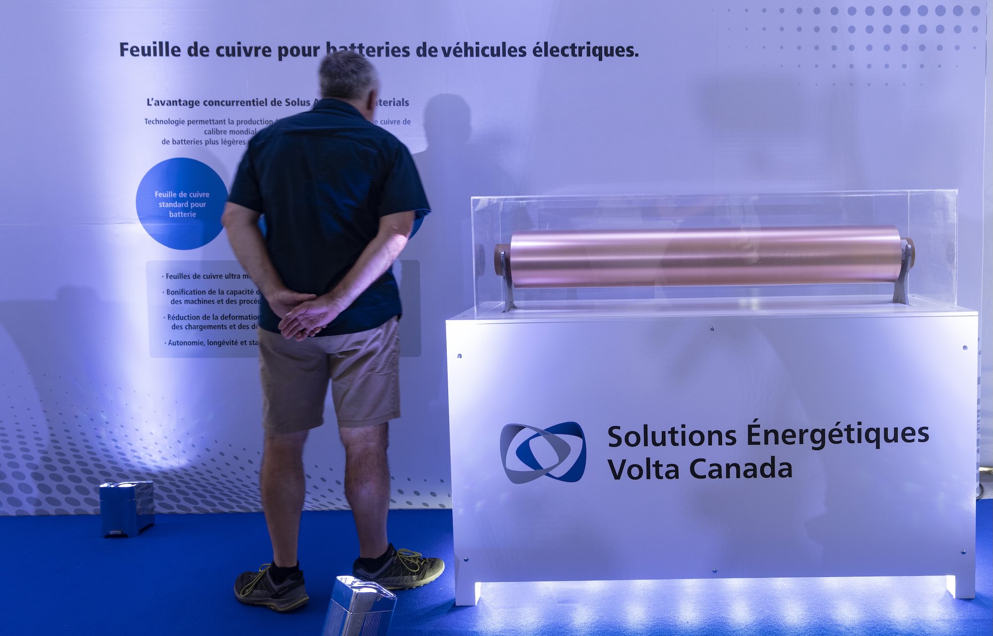 A man wearing a dark t-shirt and beige shorts stands facing a display of a sample roll of copper foil, visible in the foreground. A sign below the foil reads Solutions énergétiques Volta Canada.