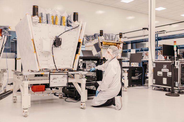 A person wearing a lab coat, mask, blue vinyl gloves and head covering kneels in front of satellite machinery.