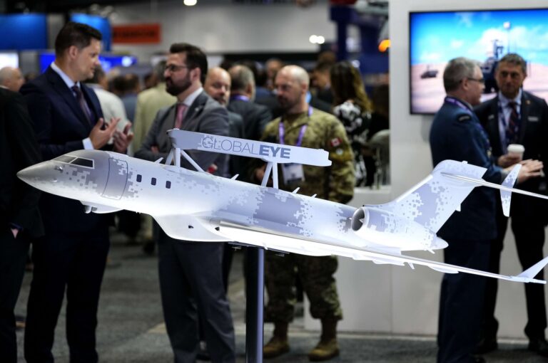A closeup of a scale model of a jet covered in pixellated camouflage, with sensor equipment attached to the top of its fuselage. There are civilians and uniformed military personnel milling in the background.
