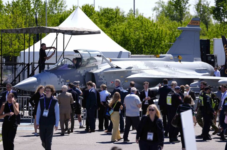 A wide shot of a lineup beside a fighter jet with the word "Gripen" printed on its tail.