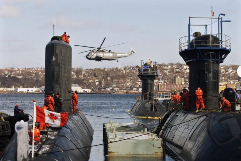 Three submarines docked side by side, crew in orange suits atop them. A helicopter hovers above, a Canadian flag visible. Coastal view of Halifax in the background.