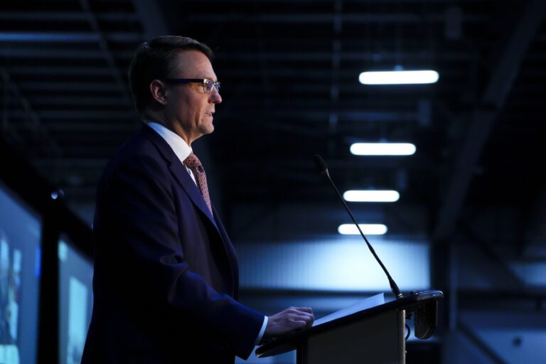 A silhouette of a man wearing a suit, tie and glasses standing at a podium with a microphone. The background is dark and overhead lights are seen in the background.