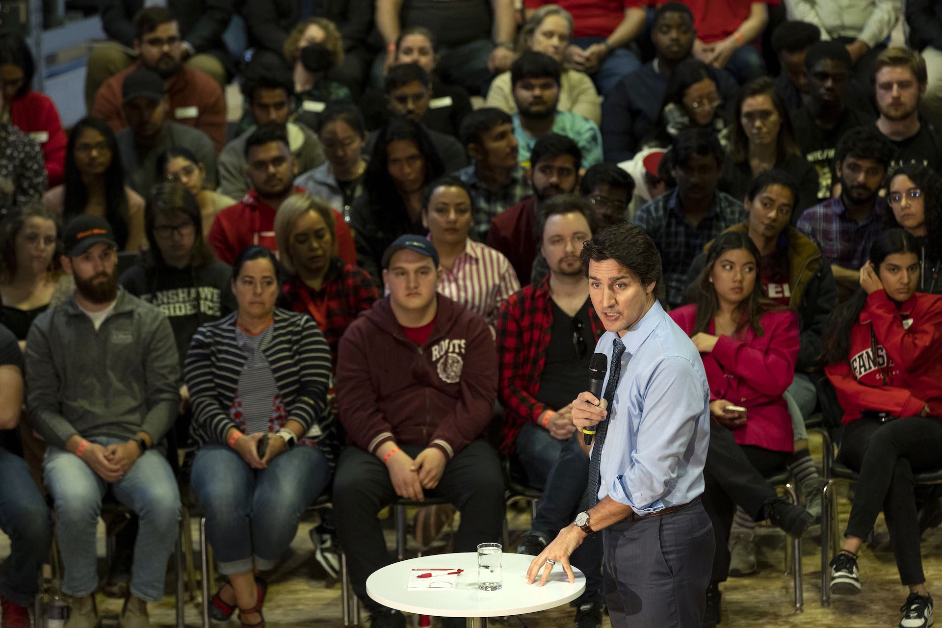A high-angle shot of Justin Trudeau speaking to a crowd of college students. He is standing before a round, white side table, and wearing a tie and a blue dress shirt with the sleeves rolled up.