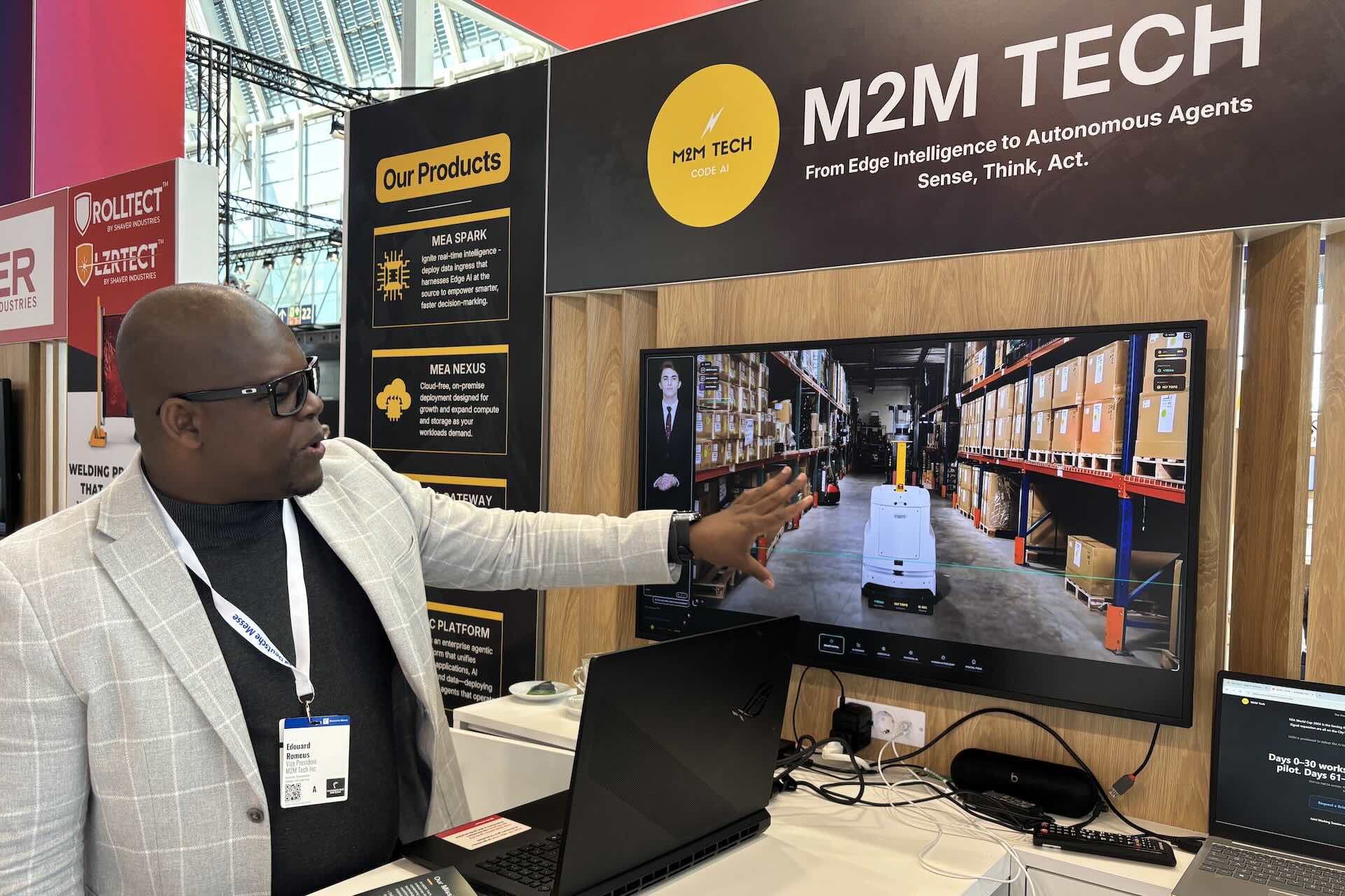 A man in a white blazer gestures towards a screen displaying a warehouse scene with a robot. A sign with "M2M Tech" is at the top of the booth.