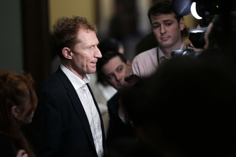 A high-angle shot of Marc Miller passing through a group of reporters and cameras. He is staring straight ahead and has pursed his lips.