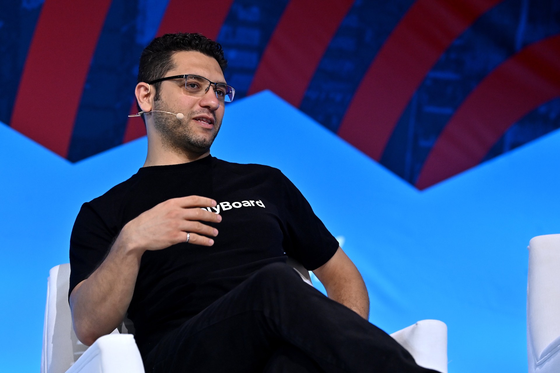 A shot of Martin Basiri onstage against a colourful backdrop, leaning back in a chair. He is wearing a black T-shirt bearing ApplyBoard's logo, and a headset mic.