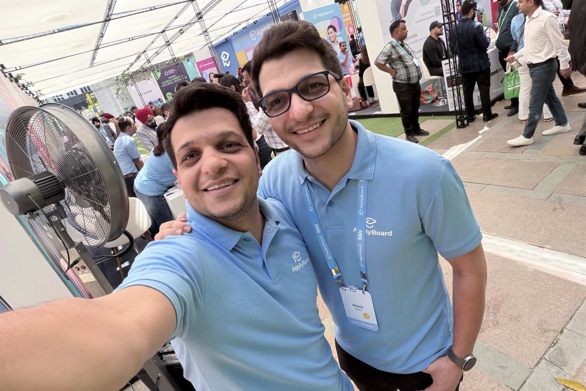 A selfie taken by Meti Basiri of him and his brother Massi inside an event pavilion. They're both wearing light blue golf shirts bearing ApplyBoard's logo.