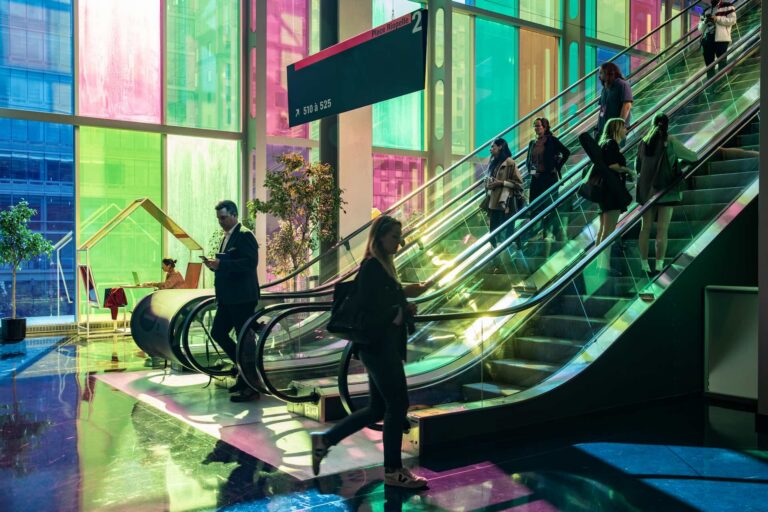 An image of the ground floor of an office building, showing two busy escalators. In the background, multicoloured glass panels are visible.