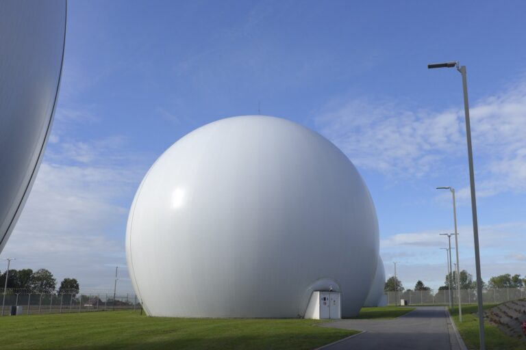 A photo of a satellite complex, with one of several giant, white spheres that house the dishes in the centre of the frame. There is blue sky and chainlink fencing in the background.
