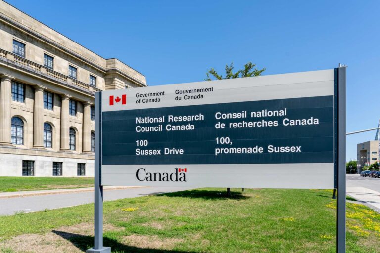 A shot of the National Research Council's headquarters in Ottawa, with its sign in the foreground. The stone building has arched windows on the ground floor and a long, third-floor balcony supported by neo-classical columns.