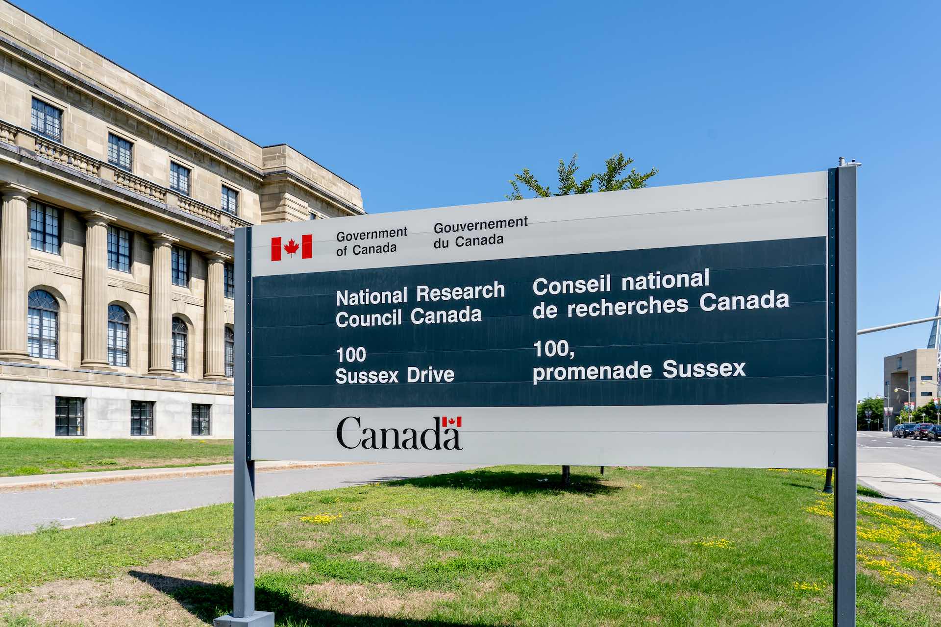 A shot of the National Research Council's headquarters in Ottawa, with its sign in the foreground. The stone building has arched windows on the ground floor and a long, third-floor balcony supported by neo-classical columns.