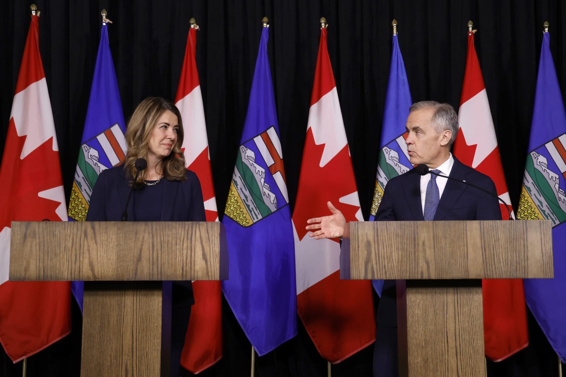 Carney and Smith standing behind identical wooden lecterns placed a couple of feet apart. There are Alberta and Canadian flags in the background.