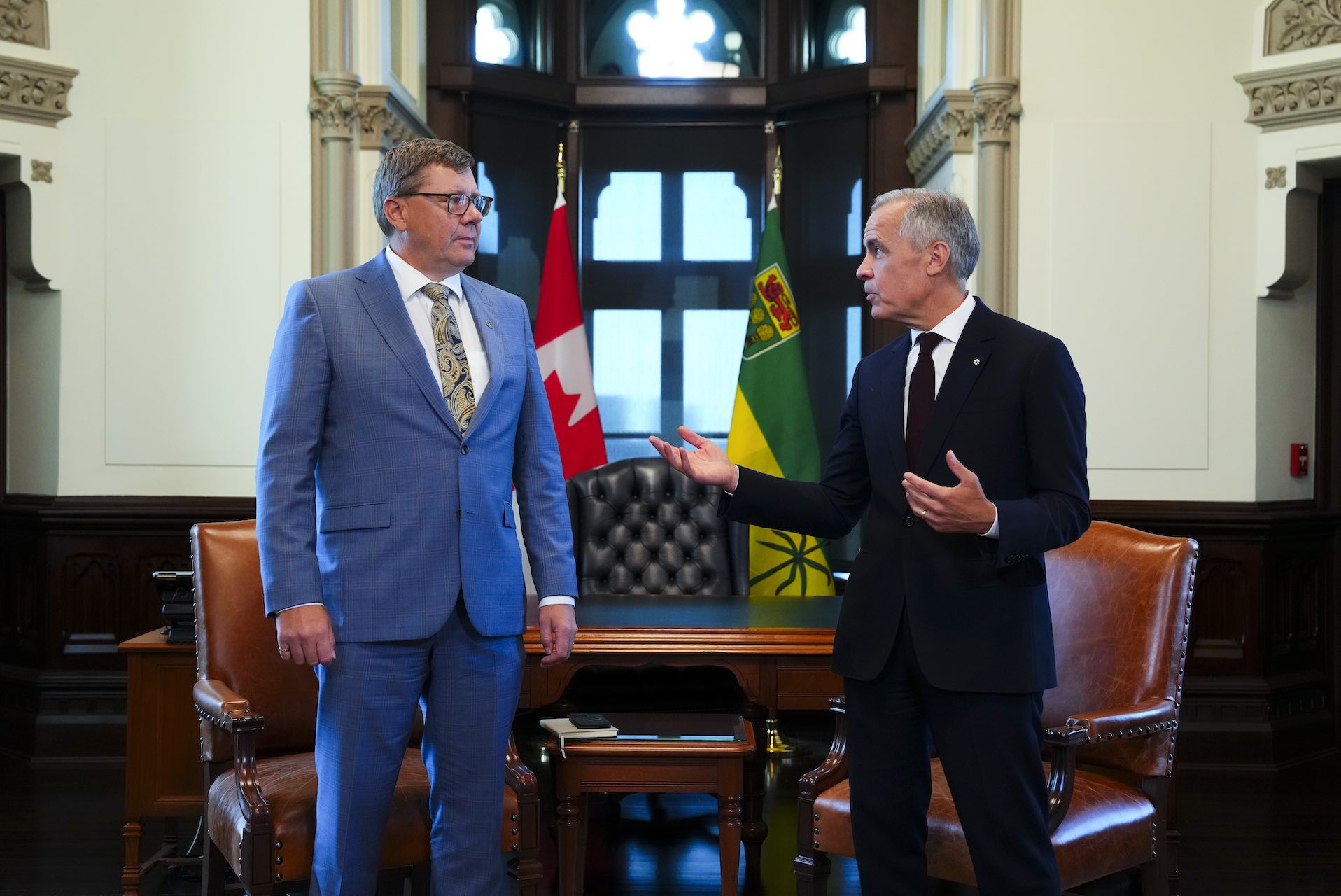 Scott Moe stands to Mark Carney's right in a room in Parliament. Carney is speaking to Moe, with hands raised slightly. There are Canadian and Saskatchewan flags in the background, behind a table with leather chairs.