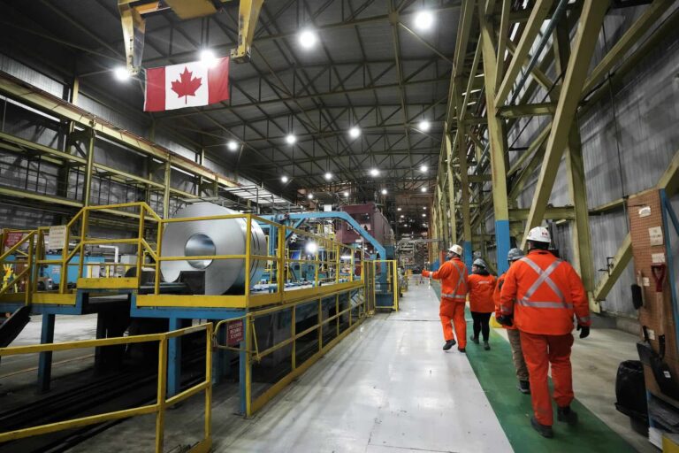 Workers in bright orange safety gear walk through a spacious industrial warehouse with machinery surrounded by yellow railings and a Canadian flag overhead.
