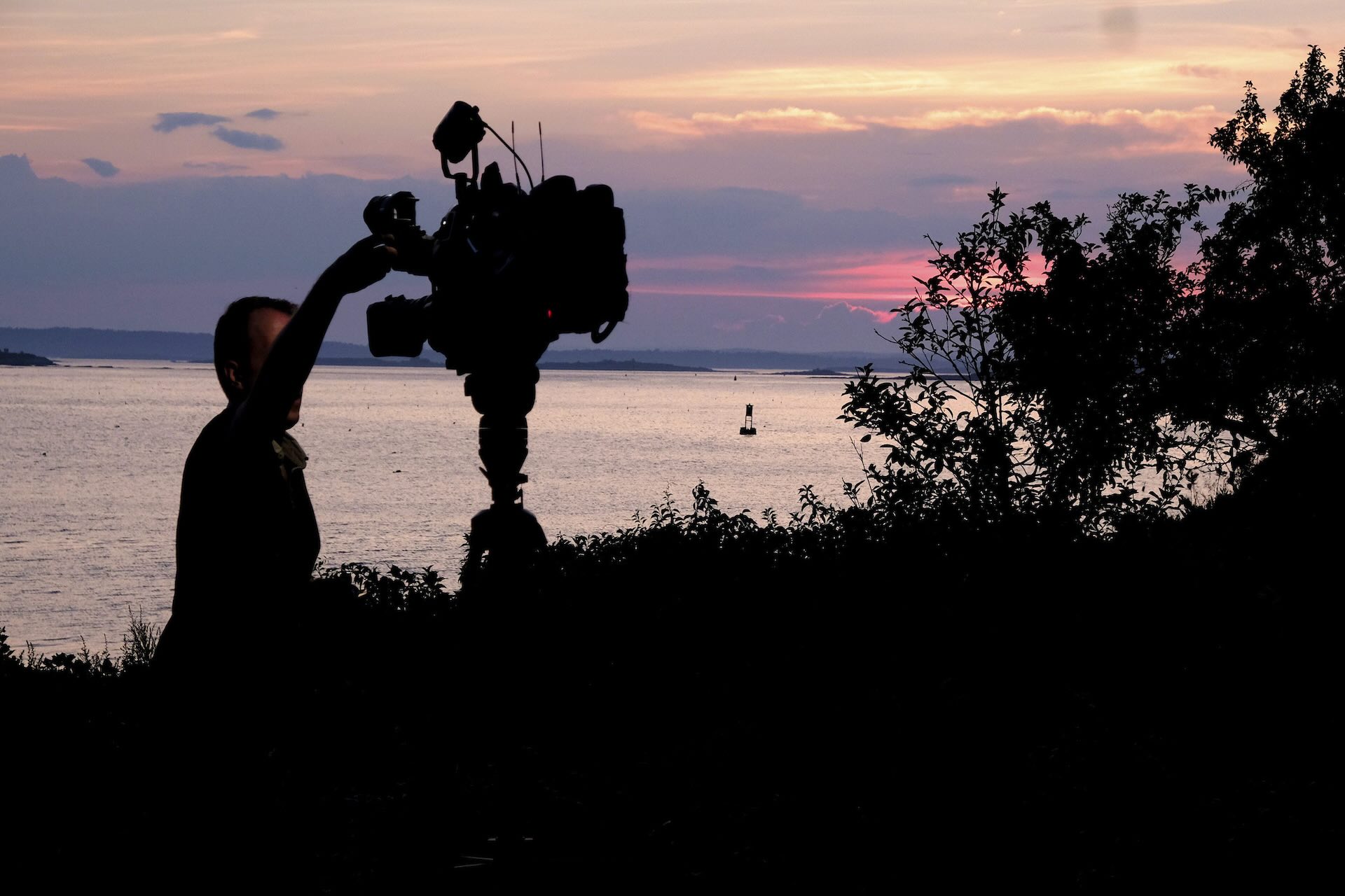 A member of a TV crew adjusts their camera while filming near the shore.