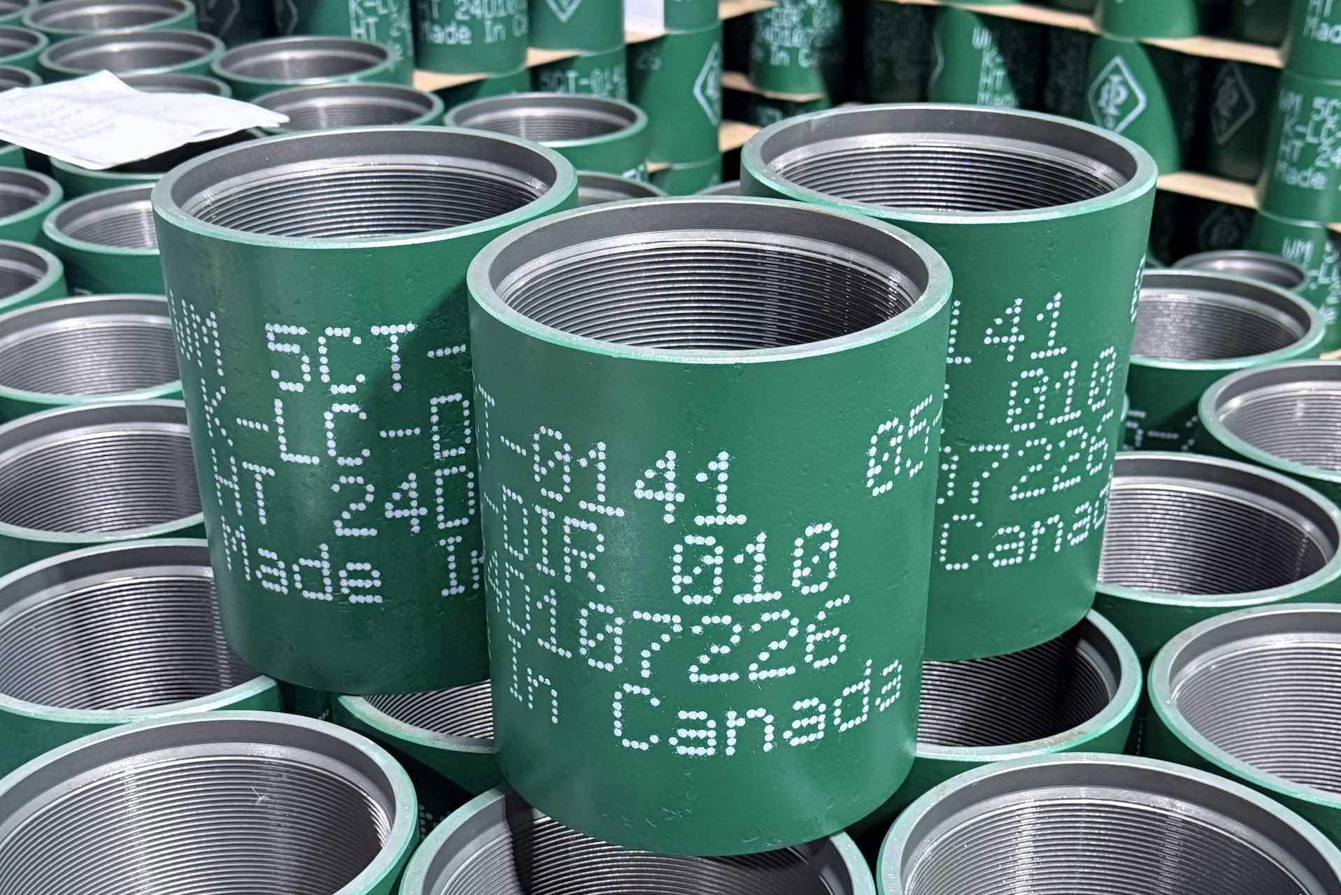 A close-up of cylindrical pipe fittings in a stack. They are green on the outside and threaded on the inside. The stamps on their exteriors reads "Made in Canada."