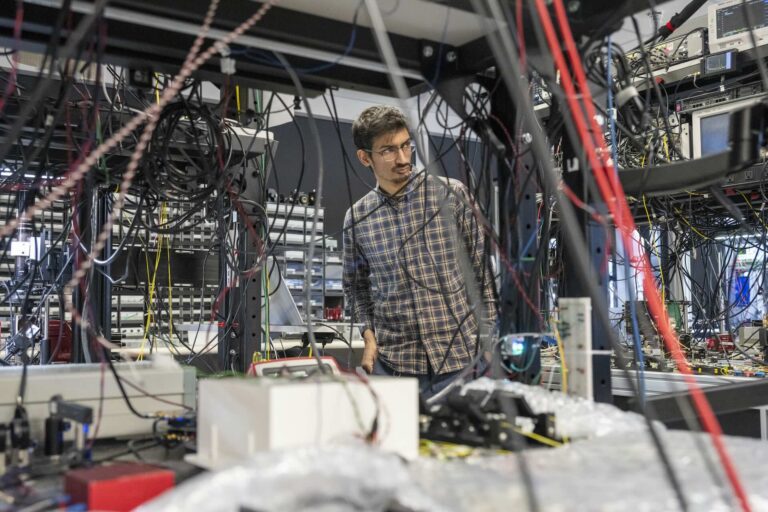 A man in a plaid button-down shirt and glasses is visible through a nest of wires in a room with shelves laden with servers, computer monitors and other equipment.