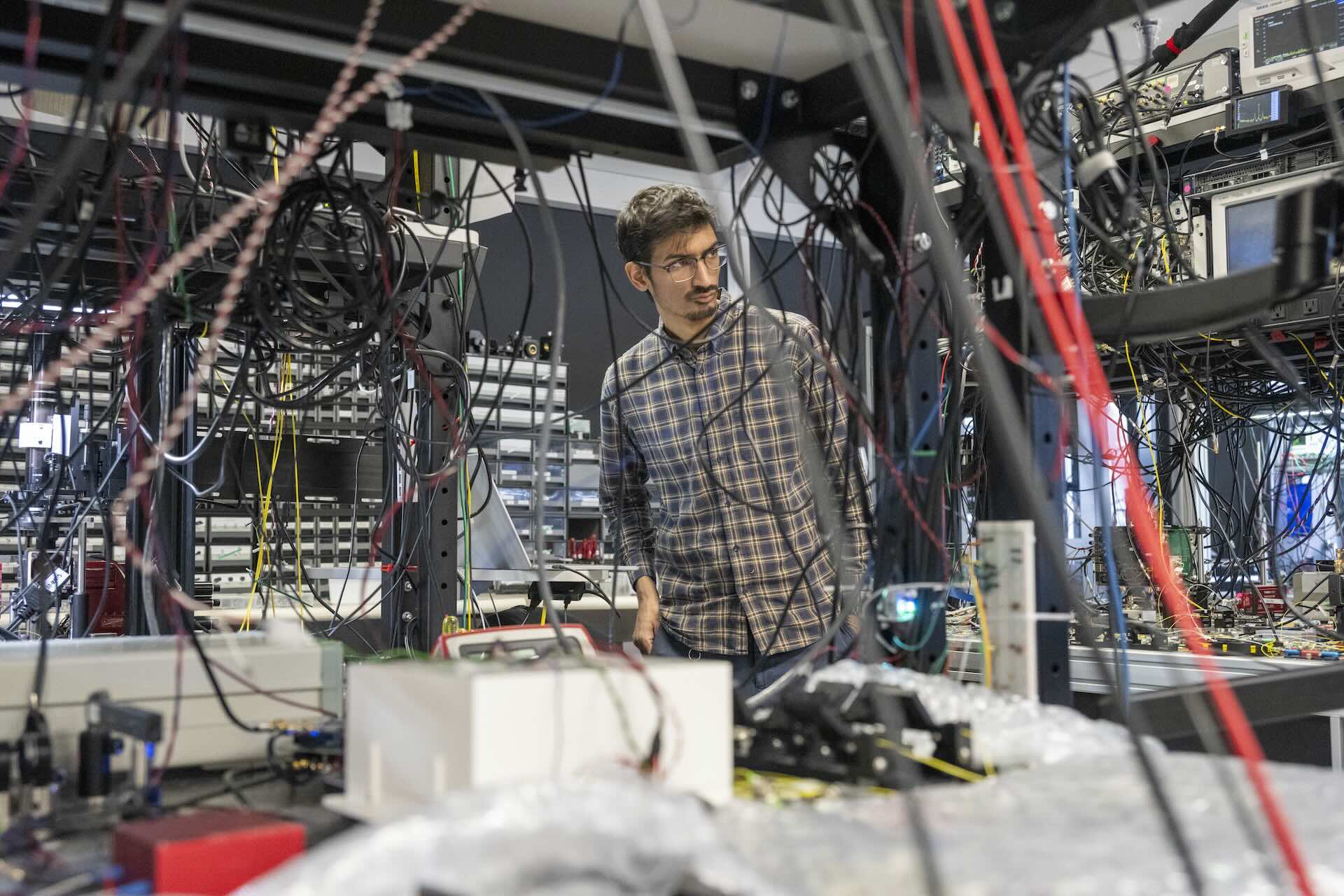 A man in a plaid button-down shirt and glasses is visible through a nest of wires in a room with shelves laden with servers, computer monitors and other equipment.